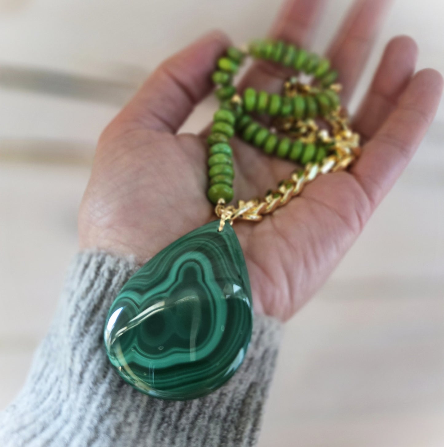 Hand holding a green malachite pendant necklace with a gold chain on a neutral background